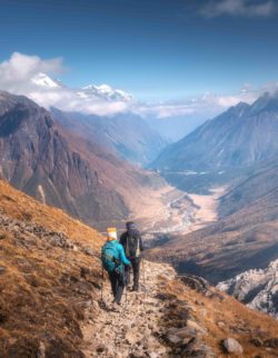 Walking people on the mountain trail at sunny day. Landscape with man and woman with backpacks, mountain valley, rocks with snowy peak in clouds, blue sky. Active people. Travel in Nepal. Trekking